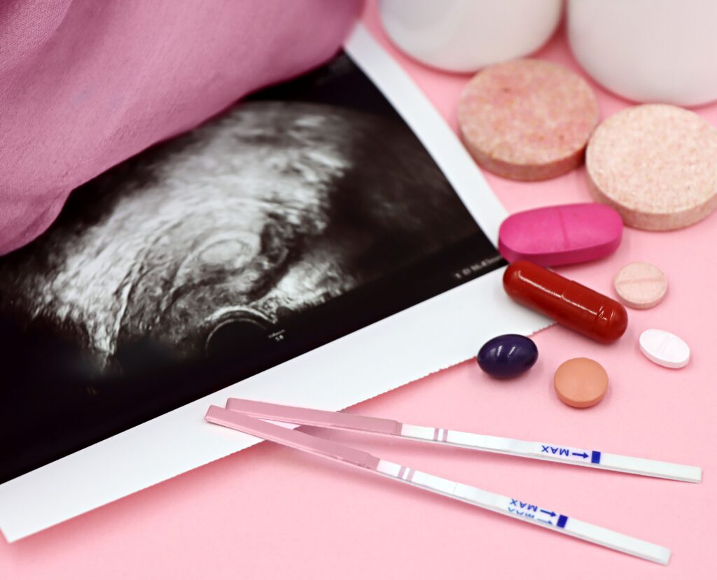 A closeup of pregnancy tests and an ultrasound with supplements on a pink background.